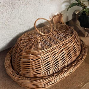 Op de afbeelding: Ovale rieten mand met een deksel en een handvat. De mand is gemaakt van geweven bruin riet en heeft een natuurlijke, rustieke uitstraling. Het deksel past goed op de basis en het handvat is een eenvoudige lus.