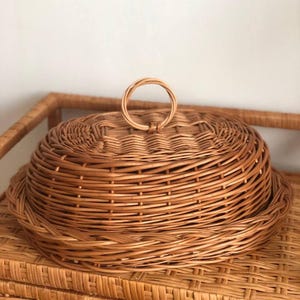 Puede incluir: Una cesta redonda de mimbre claro con tapa a juego y asa circular. La cesta y la tapa son de color marr&oacute;n claro y parecen estar hechas de materiales naturales. La cesta est&aacute; sobre una mesa de mimbre.
