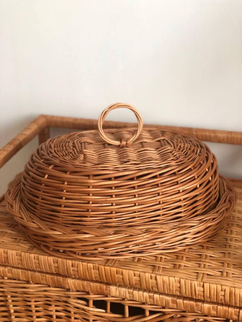 Puede incluir: Una cesta redonda de mimbre claro con tapa a juego y asa circular. La cesta y la tapa son de color marr&oacute;n claro y parecen estar hechas de materiales naturales. La cesta est&aacute; sobre una mesa de mimbre.