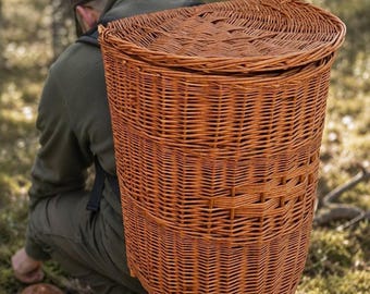 May include: A tall, cylindrical woven wicker basket with a flat, hinged lid, designed to be worn as a backpack. The basket features a natural light brown finish and a sturdy, tightly woven structure, providing a deep storage compartment.