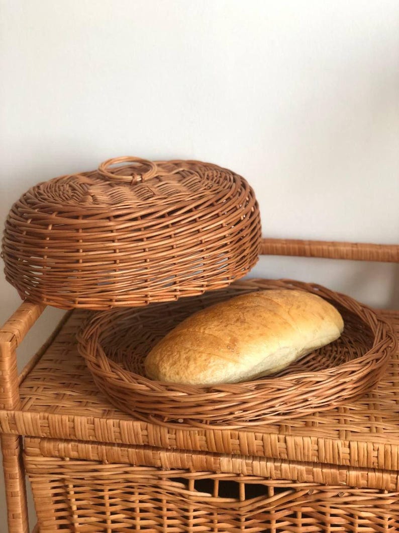 Puede incluir: Una cesta de pan redonda de mimbre con tapa y una hogaza de pan. La cesta y la tapa son de mimbre tejido. El pan es de color marr&oacute;n dorado y est&aacute; en la cesta abierta. La cesta est&aacute; sobre una estanter&iacute;a de mimbre.