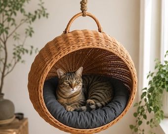 May include: A round, light brown woven wicker hanging cat bed suspended by a thick rope. The interior features a dark gray circular cushion where a brown tabby cat is resting. The structure has a curved handle at the top for hanging support.