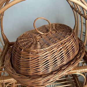 Puede incluir: Una cesta de mimbre tejida de color marr&oacute;n natural, con tapa y asa. La cesta, de forma ovalada, se encuentra sobre una estanter&iacute;a de mimbre, con un respaldo curvo.
