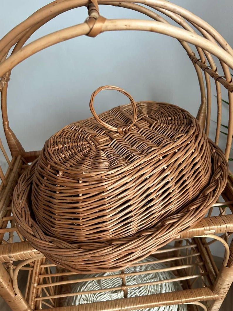 Puede incluir: Una cesta de mimbre tejida de color marr&oacute;n natural, con tapa y asa. La cesta, de forma ovalada, se encuentra sobre una estanter&iacute;a de mimbre, con un respaldo curvo.