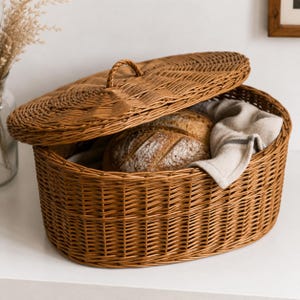 May include: A brown wicker bread basket with a lid, slightly ajar to show two loaves of bread and a linen cloth. The oval basket has a handle on the lid. The basket is designed for storing bread. The basket is made of woven wicker.