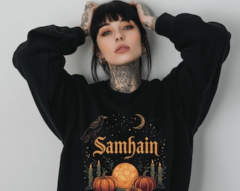 May include: Black sweatshirt featuring a graphic design with a raven, moon, pumpkins, and candles. The word "Samhain" is written in gold script. The design has a celestial and autumnal theme.