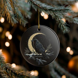May include: Black ceramic Christmas ornament featuring a raven perched on a snow-covered branch, set against a crescent moon and starry night sky. The ornament hangs from a gold string, perfect for holiday decor.