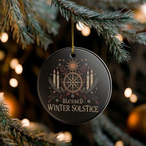May include: Black round Christmas ornament with gold string, featuring a sun, candles, and the text "Blessed Winter Solstice." The ornament is decorated with botanical and celestial elements.