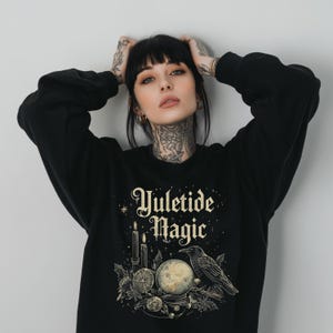 May include: Black sweatshirt with the text "Yuletide Magic" in a vintage font. The design includes a candle, a crystal ball, a raven, and other mystical elements. The sweatshirt is a cozy, long-sleeved garment.