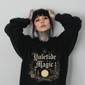 May include: Black sweatshirt with the words "Yuletide Magic" in gold script. The design includes a raven, candles, and a crystal ball. The sweatshirt is a crew neck style.