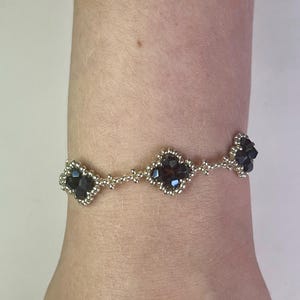 May include: A beaded bracelet featuring three dark, possibly black, floral-shaped clusters. The bracelet is made of small, silver-colored beads and is worn on a wrist. The bracelet's design is symmetrical, with the floral clusters evenly spaced.