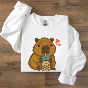 Könnte beinhalten: Weißes Sweatshirt mit einem Cartoon-Wasserschwein, das Bubble Tea trinkt. Das Wasserschwein ist braun mit rosa Wangen und hält einen teal-farbenen und braunen Bubble Tea-Becher. Zwei rote Herzen schweben über dem Wasserschwein. Das Sweatshirt hat lange Ärmel.