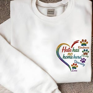 Könnte beinhalten: Weißes Sweatshirt mit Rundhalsausschnitt und farbenfrohem Herzdesign. Das Herz zeigt den Text "Hate has no home here" und Pfotenabdrücke mit den Worten "Kindness", "Equality", "Diversity" und "Love".