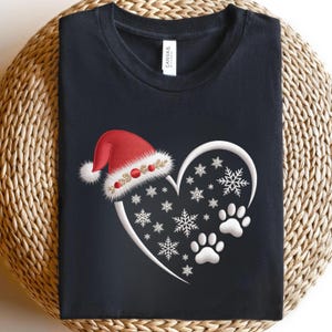 Könnte beinhalten: Schwarzes T-Shirt mit einem weißen Herzdesign, Schneeflocken und Pfotenabdrücken. Ein roter Weihnachtsmannhut mit weißer Bordüre und goldenen Akzenten befindet sich oben auf dem Herzen. Das T-Shirt ist gefaltet.
