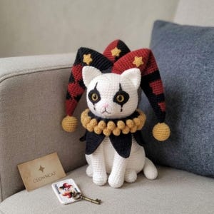 May include: A white crocheted cat doll wearing a jester hat with red and black diamond patterns, yellow stars, and pom-poms. It has a yellow ruffled collar and black eye markings. Beside it is a card labeled CLOWN CAT and a small key keychain.