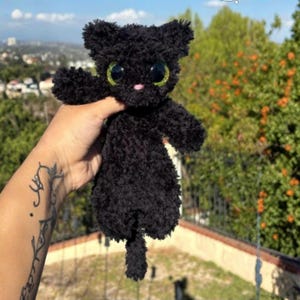 May include: A small, fuzzy black plush cat toy with large, round, bright green eyes and a tiny pink stitched nose. The stuffed animal features a textured, yarn-like material throughout its body, including pointed ears, two arms, and a tail.