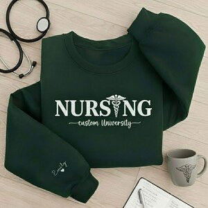 Könnte beinhalten: Dunkelgrünes Sweatshirt mit dem Wort "NURSING" in weißen Blockbuchstaben und einem Caduceus-Symbol. Der Text "custom University" steht unter dem Wort "NURSING". Eine passende Tasse mit einem Caduceus-Symbol ist ebenfalls zu sehen.