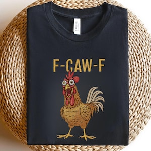 Könnte beinhalten: Schwarzes T-Shirt mit einem Cartoon-Hahn-Motiv und dem Text "F-CAW-F" in Gold. Der Hahn hat große Augen und einen offenen Schnabel, mit rotem Kamm und Kehllappen. Das T-Shirt ist aus weichem Material.