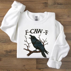 May include: White sweatshirt with the text "F-CAW-F" above a black raven perched on a brown branch. The raven is depicted with its beak open, and the design includes bare tree branches.