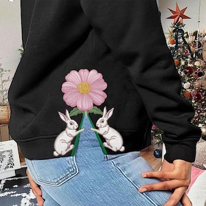 Puede incluir: Sudadera negra con una flor rosa y dos conejos blancos. Los conejos flanquean un triángulo verde. La sudadera se combina con vaqueros azules. Fondo con un árbol de Navidad.