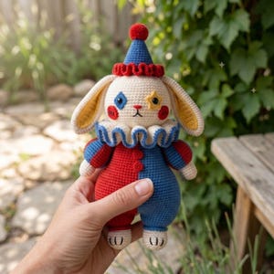 Könnte beinhalten: Handgefertigte Häkelkaninchenpuppe als Clown. Das Kaninchen hat ein weißes Gesicht mit blauen und roten Akzenten, ein blau-rotes Outfit und einen Rüschenkragen. Es trägt einen blauen Hut mit einem roten Bommel.