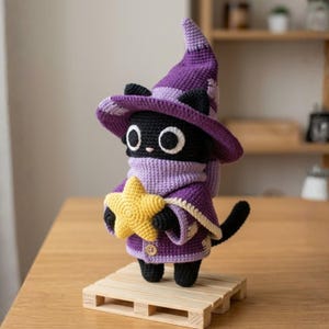 May include: A black crocheted cat figurine wearing a purple pointed hat and a matching purple cape with a light purple scarf. The cat has large white eyes with black pupils and is holding a small, textured yellow star in its front paws.