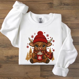 Könnte beinhalten: Weißes Sweatshirt mit einem Cartoon-Highland-Rind, das eine rote Mütze und einen Schal trägt. Das Rind hält eine Tasse heiße Schokolade mit einem Schneeflockenmuster. Goldene Weihnachtsbäume und rote Schneeflocken sind ebenfalls abgebildet.