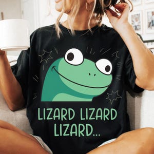 May include: Black t-shirt featuring a cartoon lizard with a green head and white eyes. The text "LIZARD LIZARD LIZARD..." is printed below the lizard graphic. The shirt is a casual style.
