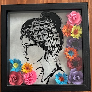 May include: Framed artwork with a black and white silhouette of a person wearing glasses, hair in a bun, filled with a library design. The frame is decorated with colourful paper flowers in pink, orange, yellow, blue, and purple.