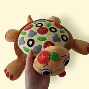 Op de afbeelding: Een pluche schildpadspeeltje met een schild in de vorm van een pizza, voorzien van vilt-toppings zoals rode tomatenschijfjes, zwarte olijfringen, groene basilicumblaadjes en grijze champignons op een gele achtergrond, met bruine ledematen.