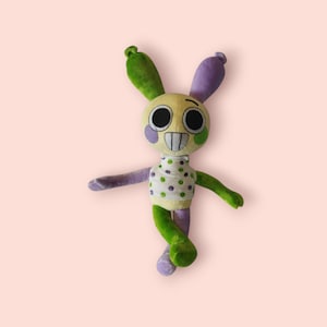 Puede incluir: Un peluche de conejo con un diseño caprichoso. El conejo presenta una cara amarilla, ojos negros grandes y una sonrisa cosida. Sus orejas son verdes y moradas, y su cuerpo es una mezcla de verde, morado y blanco con lunares. El juguete está sobre un fondo rosa.