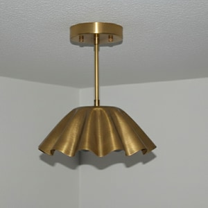 May include: A gold-colored, scalloped pendant light fixture. The light has a round ceiling mount and a slender rod connecting to the shade. The shade has a ruffled, umbrella-like design. The fixture is suspended from a white ceiling.