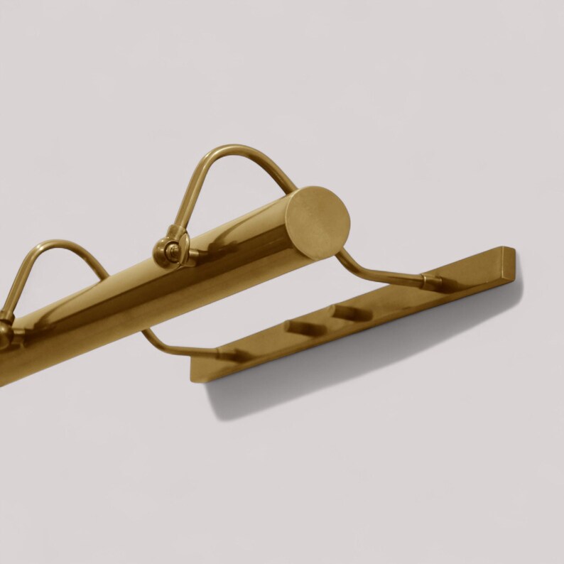 May include: A brass-toned picture light fixture. The fixture has a cylindrical main body with curved arms and a rectangular base with three small, evenly spaced hooks. The light is mounted on a white wall.