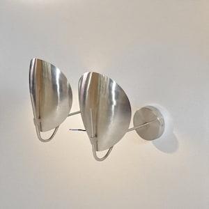 Puede incluir: Dos apliques de pared de metal cepillado. Cada aplique presenta una pantalla en forma de media luna y un brazo de soporte curvado. Los apliques están montados sobre una base circular. El cableado es visible. Diseño moderno y minimalista.