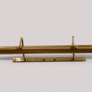 May include: A brass-colored picture light fixture with a horizontal bar and two curved arms. The fixture has a rectangular base with three small circular lights. The background is a plain white wall.