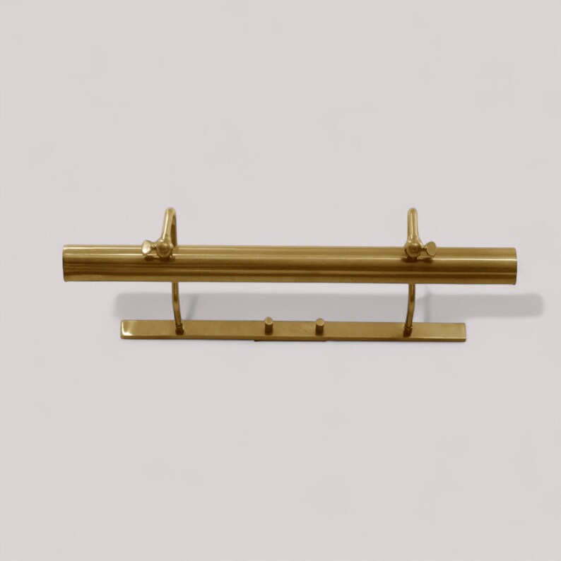 May include: A brass-colored picture light fixture. The fixture has a long, cylindrical light bar with two adjustable arms and a rectangular base. The light is designed to be mounted on a wall and is suitable for illuminating artwork.