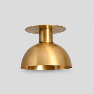 May include: A gold-colored, dome-shaped ceiling light fixture. The fixture has a circular base, a cylindrical stem, and a rounded shade. The metal has a brushed finish, giving it a warm, inviting appearance. The light is designed for indoor use.