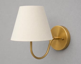 May include: A wall-mounted sconce with a cream-colored lampshade and a brass finish. The lampshade is cone-shaped, and the fixture has a curved arm connecting the shade to a circular wall plate. The sconce is designed for indoor use.