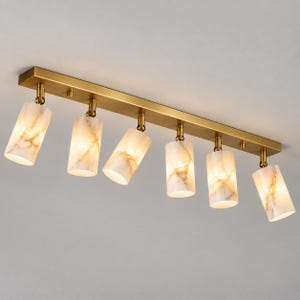 May include: A ceiling light fixture featuring a long, rectangular brass-toned metal base with six adjustable cylindrical spotlights. Each spotlight has a white, marble-patterned glass shade that emits a warm, soft glow from within the fixture.