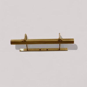 May include: A brass-colored picture light fixture. The fixture has a horizontal cylindrical bar with two curved supports and a rectangular base with three small pegs. The light is mounted on a white wall.
