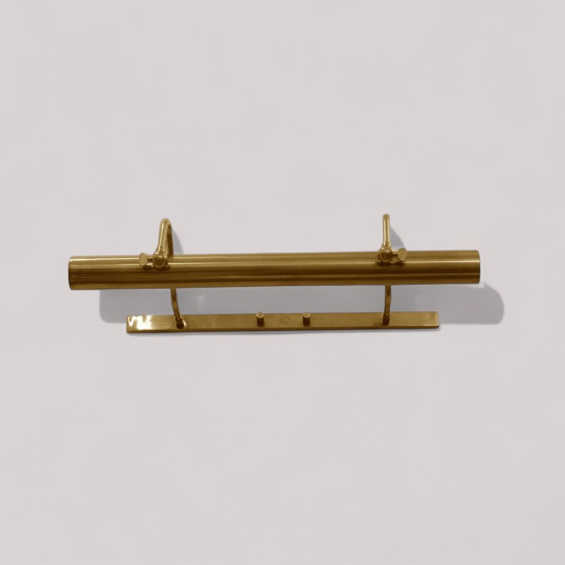 May include: A brass-colored picture light fixture. The fixture has a horizontal cylindrical bar with two curved supports and a rectangular base with three small pegs. The light is mounted on a white wall.