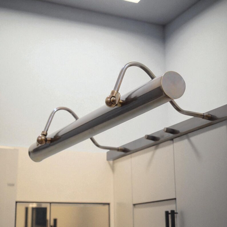 May include: A silver-toned picture light with curved arms and brass-colored accents. The cylindrical light fixture is mounted above a white cabinet. The light is designed to illuminate artwork or displays.