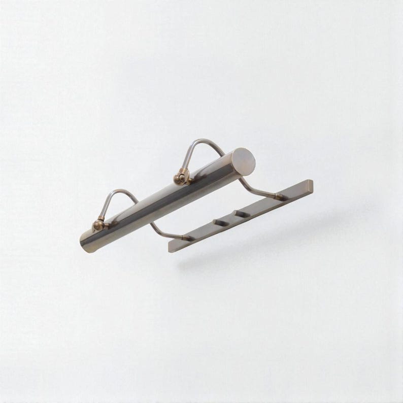 May include: A silver-toned picture light with a cylindrical body and a rectangular base. The light has two curved arms with small brass-colored accents. The base has three slots. The fixture is designed to illuminate artwork.