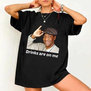May include: Black t-shirt featuring a graphic of a man in a hat with the text "Drinks are on me" in white. The graphic is a humorous image. The shirt is a casual wear item.