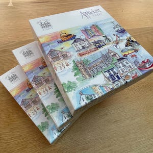 Può includere: Tre puzzle in scatola con illustrazioni colorate di una mappa della città. Le scatole bianche riportano il testo "Appledore, North Devon" e presentano un disegno dettagliato di edifici, barche e monumenti. I puzzle sono impilati su una superficie di legno.