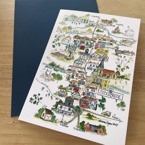 May include: A watercolor map of Stockbridge, with detailed illustrations of buildings, trees, and a river. The map is on white paper, with a dark blue envelope behind it. The text "Stockbridge" is visible.