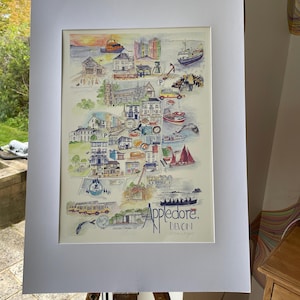 May include: A framed watercolor print of Appledore, Devon, featuring colorful illustrations of buildings, boats, and landmarks. The artwork includes the town's name in stylized lettering and is displayed on an easel.
