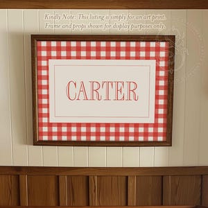 May include: A red and white gingham print with the name "CARTER" in red text. The print is framed and hanging on a wall above a wooden crib.
