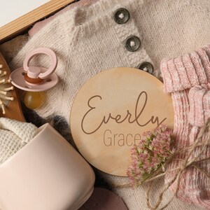 K&ouml;nnte beinhalten: Eine Holzkiste gef&uuml;llt mit Babyartikeln, darunter ein gestrickter Pullover, Socken, ein Schnuller, eine Tasse, ein geh&auml;keltes H&auml;schen, eine Haarb&uuml;rste und ein Holzschild mit dem Namen "Everly Grace".