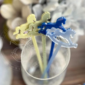 May include: Three acrylic drink stirrers in a clear glass, each featuring a rocking horse topper. The stirrers are in pale yellow, dark blue, and translucent light blue. A circular watermark with text is visible over the stirrers.